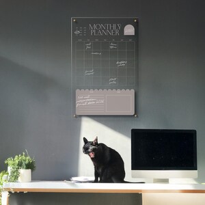May include: A clear acrylic monthly planner with gold hardware is mounted on a gray wall. The planner has a calendar grid and a note section. A black cat yawns on a white desk below the planner, next to a computer monitor and a potted plant.