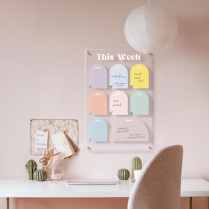 May include: A clear acrylic weekly planner with pastel-colored arched sections for each day. The planner has handwritten notes and the words "This Week" at the top. A white paper lantern hangs above the planner, and a desk with decorative items is below.