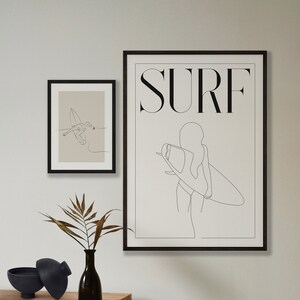 May include: Two black framed prints with line drawings of a person surfing. The larger print has the word "SURF" in bold letters at the top. The smaller print shows a person surfing a wave.