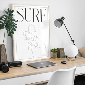 May include: A framed black and white line drawing of a person surfing with the word "SURF" in bold letters above the image. The image is hanging on a white wall above a wooden desk with a laptop, a lamp, and a moon lamp.