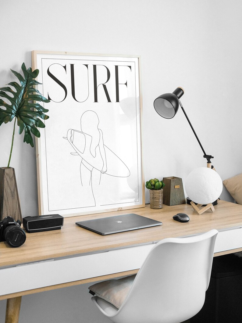 May include: A framed black and white line drawing of a person surfing with the word "SURF" in bold letters above the image. The image is hanging on a white wall above a wooden desk with a laptop, a lamp, and a moon lamp.