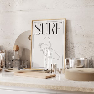 May include: A framed print with a gold frame and a white background. The print features a line drawing of a person surfing with the word "SURF" in black text at the top.