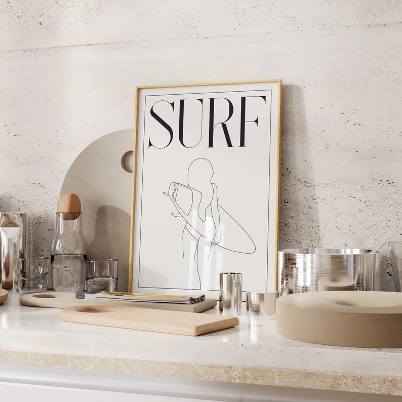 May include: A framed print with a gold frame and a white background. The print features a line drawing of a person surfing with the word "SURF" in black text at the top.