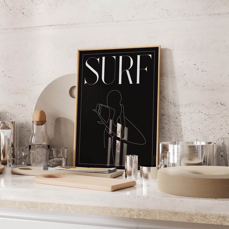 May include: A black and white illustration of a person surfing with the word "SURF" in white text on a black background. The illustration is framed in a gold frame.