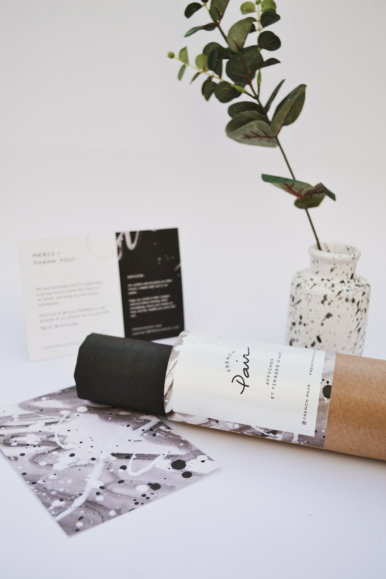May include: A rolled up poster with a white label that says "French Pair" and "Affiches et Images d'Art". The poster is sitting on a white surface with a black and white card and a small vase with a green plant.