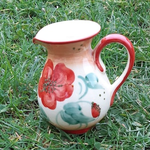 May include: A ceramic jug with a red handle and spout, hand-painted with red flowers, green leaves, and a strawberry. The jug is set on green grass. The jug is likely used for serving liquids.