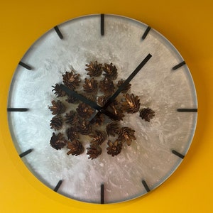 May include: A white clock face with black hands and black Roman numeral markings. The clock face is decorated with small, brown pine cones.