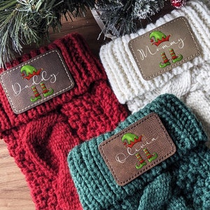 May include: Three cable-knit Christmas stockings in red, white, and green. Each stocking has a brown leather patch with a name and an elf graphic. Festive holiday decor.