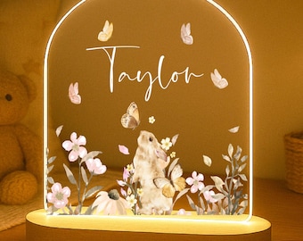May include: A decorative night light with a wooden base and an arched acrylic panel. The panel features a bunny, flowers, and butterflies, with the name "Taylor" in script. The light emits a warm glow.