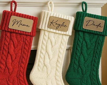 May include: Three cable-knit Christmas stockings in red, cream, and green hang from a white mantel. Each stocking has a leather patch with a name: "Mama", "Kaylee", and "Dada". A garland of frosted greenery decorates the mantel above the stockings.