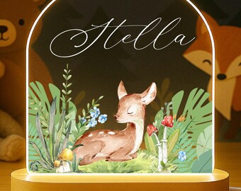 May include: A wooden-based night light with a deer illustration, the name "Stella", and an arched, illuminated frame. The background features a forest scene with a bear and fox. The light is connected to a white cord.