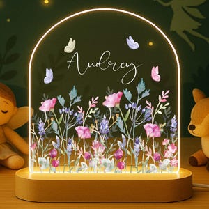 May include: A personalized night light with a floral design and the name "Audrey" in script. The light has a wooden base and an arched acrylic panel with butterflies. A fairy doll and a deer plush toy are on either side.