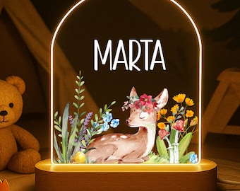 May include: A decorative night light with the name "MARTA" in white. The light features a watercolor illustration of a deer with a floral crown, surrounded by flowers and greenery. The base is wooden, and a teddy bear is in the background.