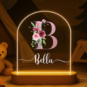 May include: A personalized night light with a floral design. The light features the letter "B" with a floral arrangement and the name "Bella" in a script font. The light has a wooden base and a warm glow, creating a cozy ambiance.