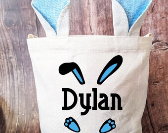 May include: White canvas Easter basket with blue bunny ears, black bunny ears, and blue paw prints. The name "Dylan" is printed in black on the front of the basket.