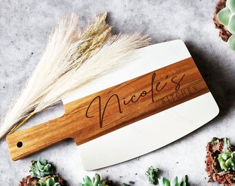 May include: A white and brown wooden cutting board with the text "Nicole's Kitchen" engraved on it. The board is surrounded by small green succulents.