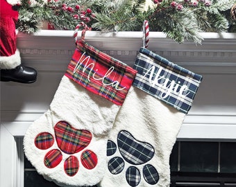 May include: Two white Christmas stockings with paw print designs. One stocking has a red and green plaid cuff with the name "Mila" embroidered on it. The other stocking has a blue and green plaid cuff with the name "Akyle" embroidered on it.