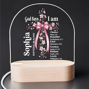 May include: A decorative light with a wooden base and a clear acrylic top. The acrylic features a cross design with pink flowers and a pink bow. Text includes "God Says I am" and "Sophia BAPTIZED 06.24.2025".