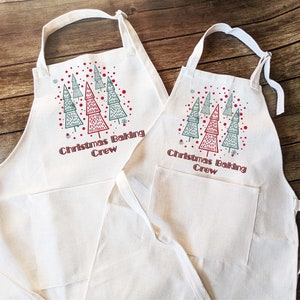 May include: Two off-white aprons with a Christmas tree design and the text "Christmas Baking Crew". The aprons have a pocket and adjustable neck strap. The Christmas tree design features red and green trees with red dots and snowflakes.
