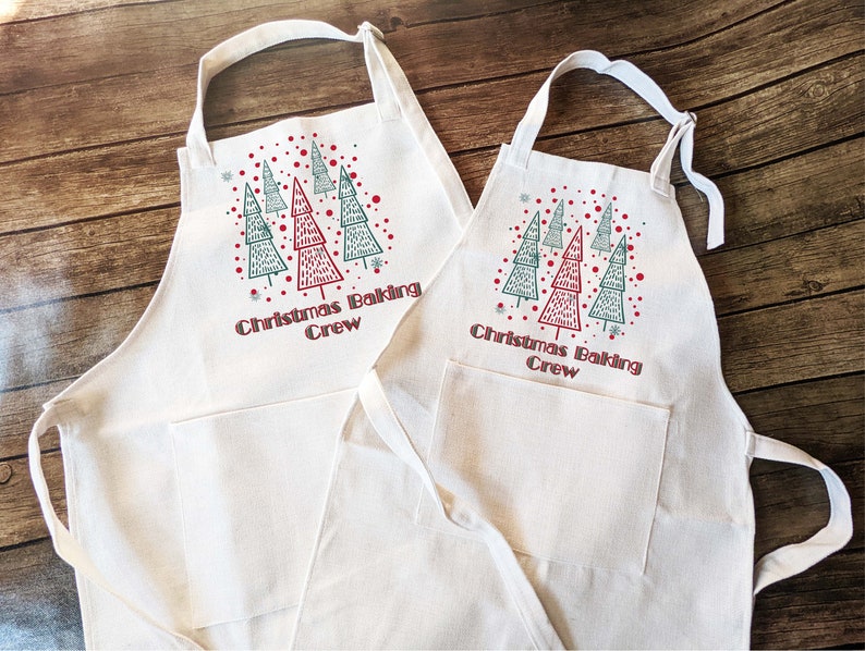 May include: Two off-white aprons with a Christmas tree design and the text "Christmas Baking Crew". The aprons have a pocket and adjustable neck strap. The Christmas tree design features red and green trees with red dots and snowflakes.