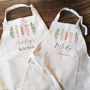 May include: Two white cotton aprons with a pocket, one with the text "Ashley's Kitchen" and the other with the text "Mila". Both aprons feature a watercolor design of baking tools and greenery.