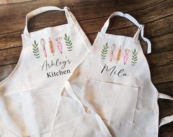 May include: Two white cotton aprons with a pocket, one with the text "Ashley's Kitchen" and the other with the text "Mila". Both aprons feature a watercolor design of baking tools and greenery.