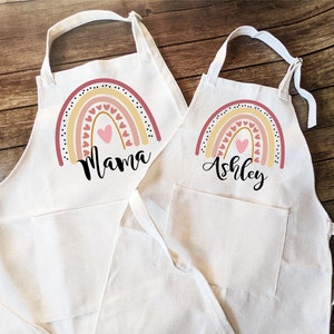 May include: Two white aprons with rainbow designs and personalized names. The larger apron says "Mama" and the smaller apron says "Ashley".