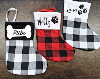 May include: Three personalized Christmas stockings with buffalo plaid fabric. The stockings are decorated with the names "Mila", "Molly", and "Luna" and paw prints.