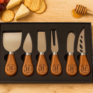 May include: A set of six cheese knives with wooden handles, each engraved with the letter "H" inside a wreath. The knives are displayed in a black box, alongside grapes, crackers, cheese, and a honey dipper.