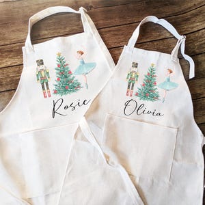 May include: Two white cotton aprons with a pocket, featuring a Nutcracker and ballerina design with a Christmas tree. The aprons are personalized with the names "Rosie" and "Olivia".