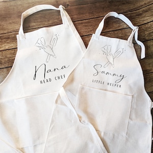 May include: Two white cotton aprons with black text and a simple illustration of a piping bag and spatula. The larger apron says "Nana Head Chef" and the smaller apron says "Sammy Little Helper".