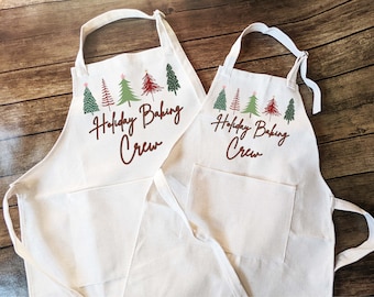 May include: Two white aprons with a pocket, featuring a line of green, red, and brown Christmas trees with a star on top. The text "Holiday Baking Crew" is written in brown cursive.
