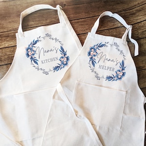 May include: Two white cotton aprons with blue floral wreaths and text. The larger apron says "Nana's Kitchen" and the smaller apron says "Nana's Helper".