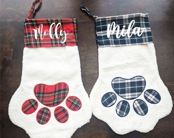 May include: Two white Christmas stockings with paw print designs. One stocking has a red and black plaid paw print and the name "Molly" on the cuff. The other stocking has a blue and white plaid paw print and the name "Mila" on the cuff.