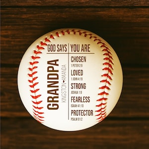 May include: A white baseball with red stitching and text that reads "GOD SAYS YOU ARE" and "GRANDPA". Additional text includes "CHOSEN", "LOVED", "STRONG", "FEARLESS", and "PROTECTOR". The baseball has the names "KINGSTON - AMANDA".