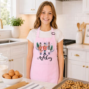 May include: A light pink apron featuring a graphic of three nutcracker figures surrounded by green pine branches and snowflakes. The name Ashley is printed in black cursive script below the nutcrackers. The apron has a neck strap and a front pocket.