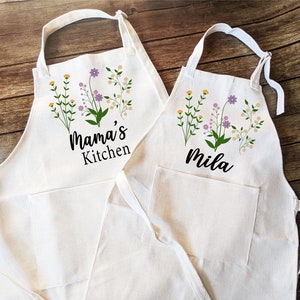May include: Two white cotton aprons with floral designs and text. The larger apron says "Mama's Kitchen" and the smaller apron says "Mila".