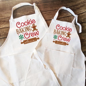 May include: Two white aprons with a pocket, featuring a gingerbread man graphic and the text "Cookie Baking Crew" with a snowflake and rolling pin. The aprons are personalized with the names "Anna" and "Linda".