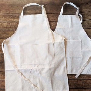 May include: Two white cotton aprons with pockets. The larger apron has adjustable straps and a longer length. The smaller apron has shorter straps and a shorter length.