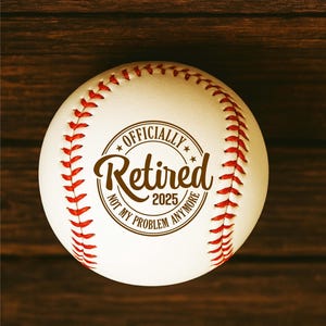 May include: A white baseball with red stitching and the text "OFFICIALLY Retired 2025 NOT MY PROBLEM ANYMORE" in a brown circular design. The baseball is set against a dark wood grain background.