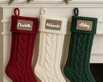 May include: Three cable-knit Christmas stockings in red, cream, and green hang from a white mantel. Each stocking has a leather patch with a name: "Charlotte," "Melanie," and "Abby." The mantel is decorated with greenery, pinecones, and ornaments.