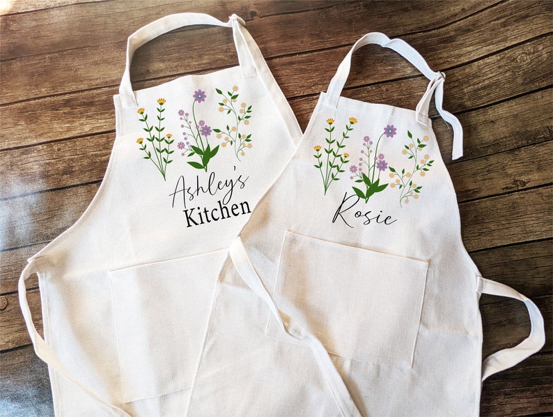 May include: Two white cotton aprons with floral designs and personalized text. The larger apron says "Ashley's Kitchen" and the smaller apron says "Rosie".