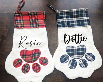 May include: Two white Christmas stockings with paw print designs. One stocking has a red and green plaid cuff with the name "Rosie" and a red paw print. The other stocking has a blue and white plaid cuff with the name "Dottie" and a blue paw print.