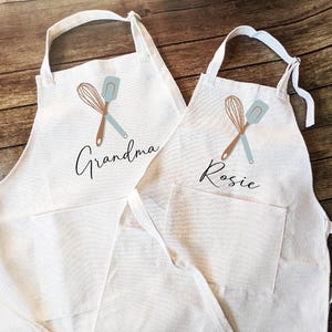 May include: Two off-white aprons with pockets, each featuring a crossed whisk and spatula design in brown and blue. One apron says "Grandma" and the other says "Rosie" in black script. The aprons are on a wooden surface.