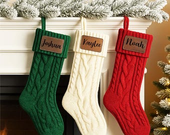 May include: Three cable-knit Christmas stockings in green, cream, and red hang from a white mantel. Each stocking has a brown leather patch with a name: Joshua, Kaylee, and Noah. The stockings are decorated with a snowy garland.