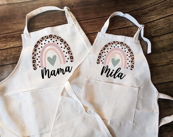 May include: Two white cotton aprons with a rainbow design and a heart. The larger apron says "Mama" and the smaller apron says "Mila".
