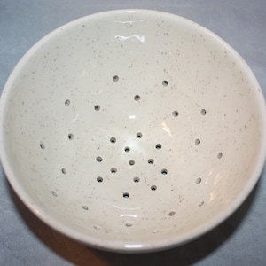 May include: A white ceramic colander with a speckled finish and a pattern of holes in the bottom.