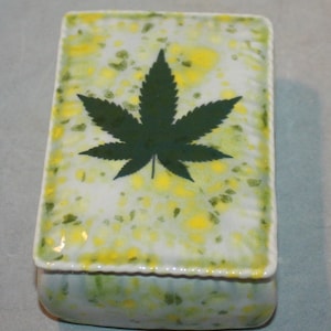 May include: A white ceramic box with a green marijuana leaf design. The box has a speckled yellow and green glaze.