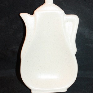 May include: A cream-colored ceramic teapot-shaped spoon rest. The spoon rest has a speckled finish and a handle-like design on the side. The lid has a small knob on top. The background is black.