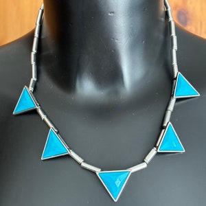 Puede incluir: Un collar de plata con una cadena de pequeños cilindros de plata y cinco colgantes triangulares turquesa.